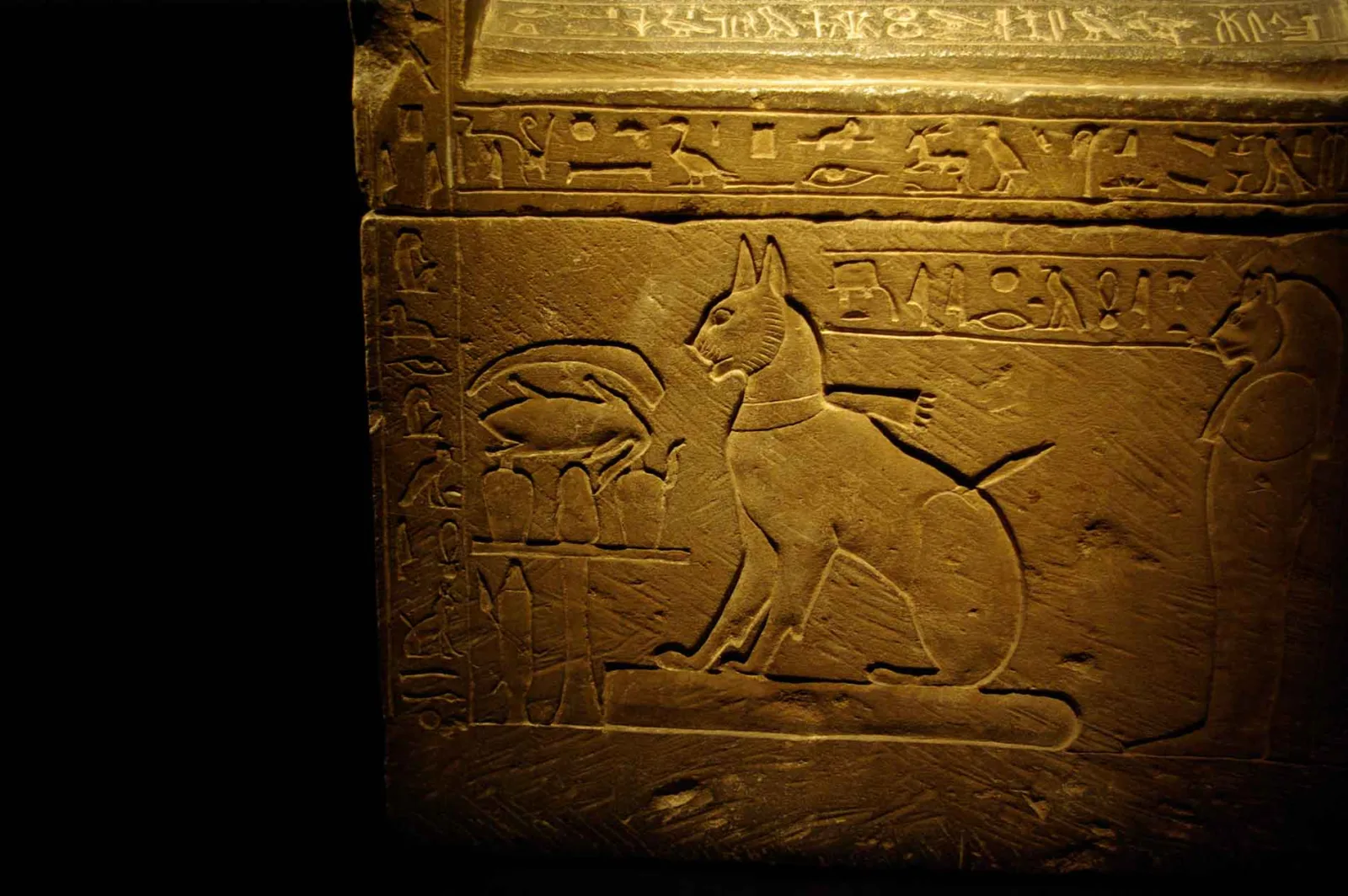 Basreliefschnitzerei einer sitzenden Katzenfigur mit spitzen Ohren und Schal. Sie sitzt vor einem Opfertisch. Hieroglyphen rahmen die Szene ein.